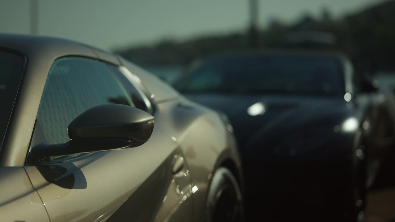 Moderna auto sportiva grigia parcheggiata, che mostra lo specchietto laterale e il finestrino, con un'auto nera sfocata sullo sfondo.
