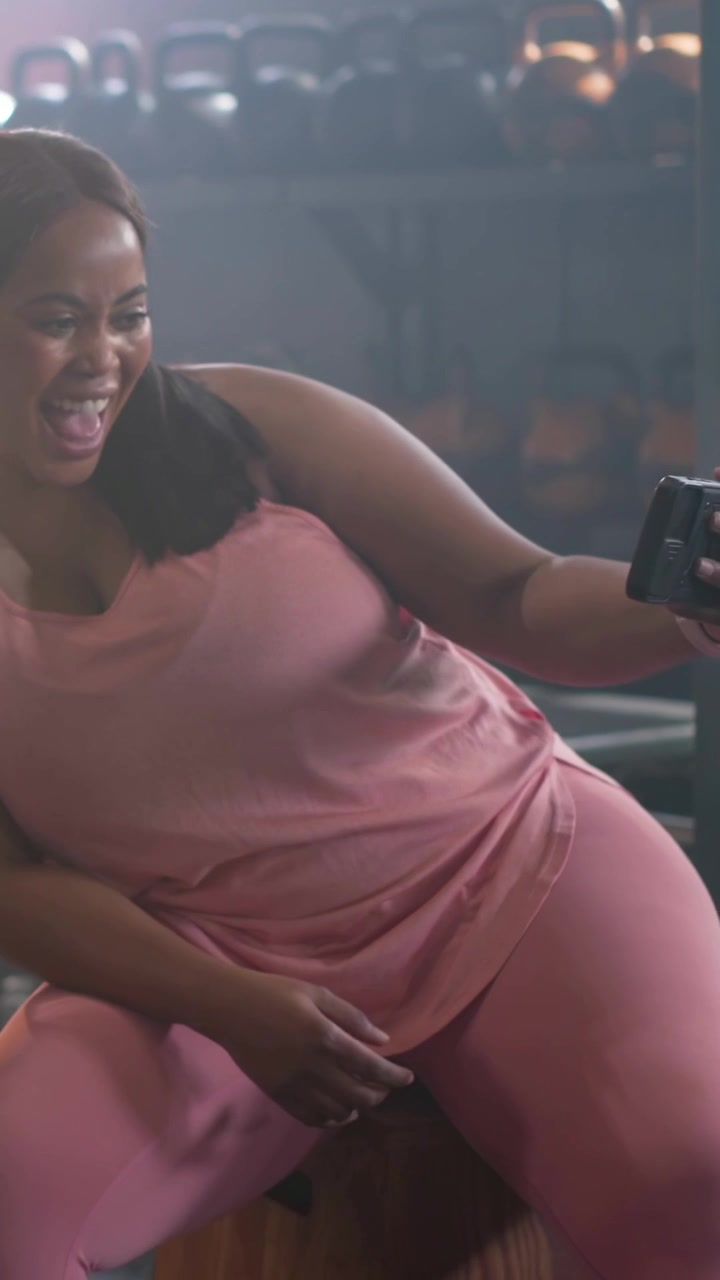 Vídeo vertical: Reagindo a um zumbido, mulher de roupa de ginástica rosa rindo, mostrando o telefone em uma caixa pliométrica na academia