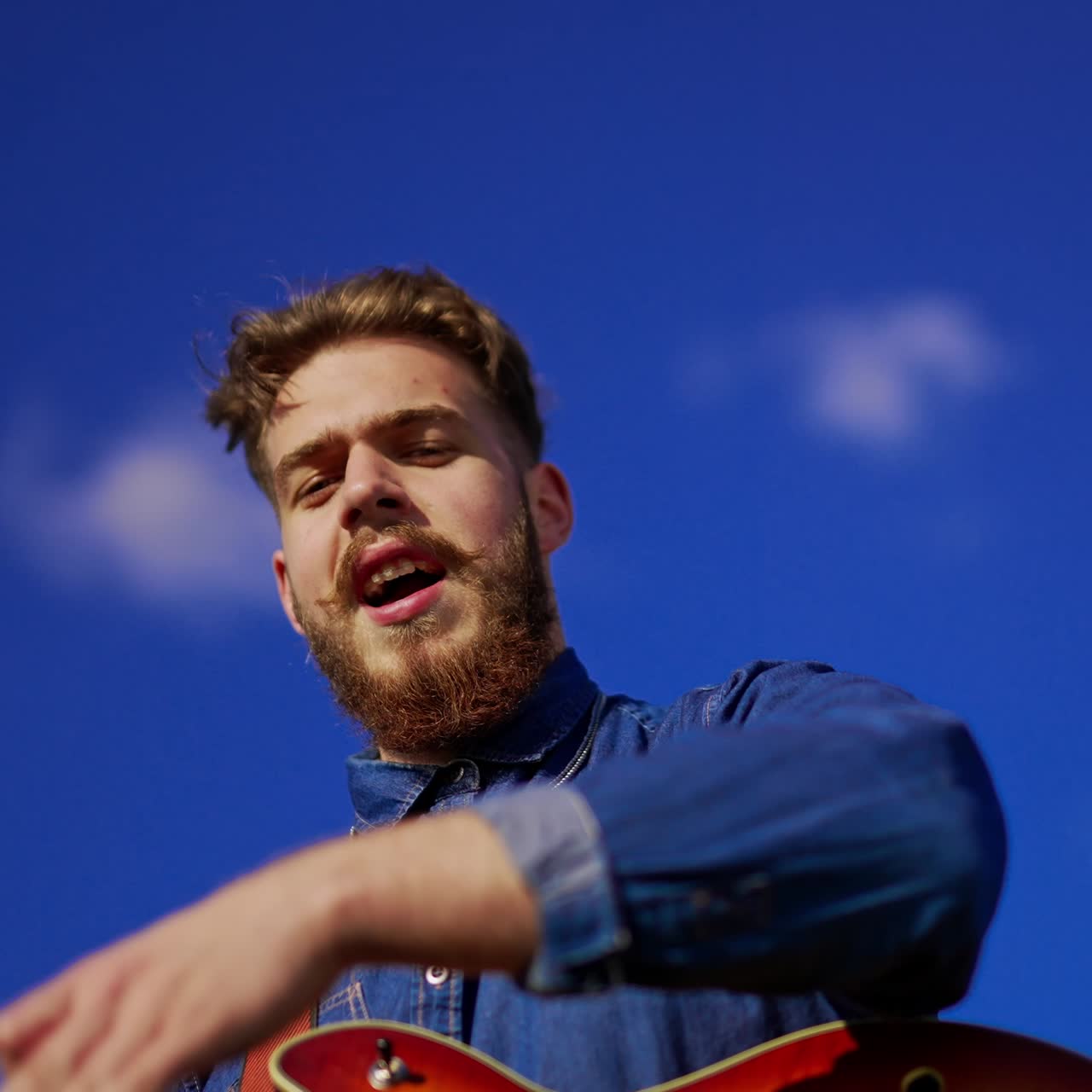 Dark-haired Caucasian man with guitar on strap dancing with his hands and singing. Close up. Low angle view. Blue sky at backdrop