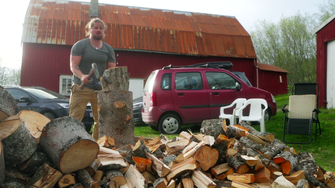 Young Man chopping wood into firewood logs outdoors beside a rustic cottage, vehicles, and chairs in a rural countryside setting, illustrating traditional manual work and lifestyle