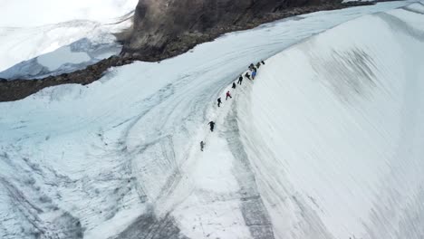 aerial drone footage orbiting and spinning around a group of people who are hiking and walking up white snow covered black colored kebnekaise mountain in sweden during a cloudy day. steep slopes seen.