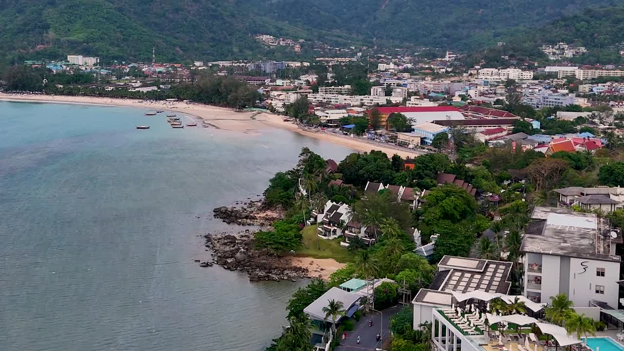 Aerial view of a coastal area featuring buildings, lush greenery, and a sandy beach under a clear sky.