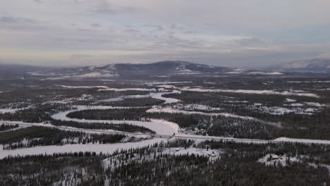 A dramatic aerial shot captures the Takhini River winding through Ibex Valley in Yukon Territory. Dense fir forests line the river, all blanketed in snow, showcasing raw winter beauty.