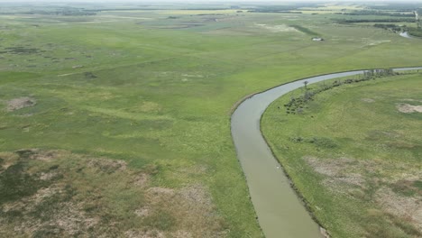 Aerial looking down at a large winding river surrounded by marsh and farm fields. A large white pelican is flying above the river.