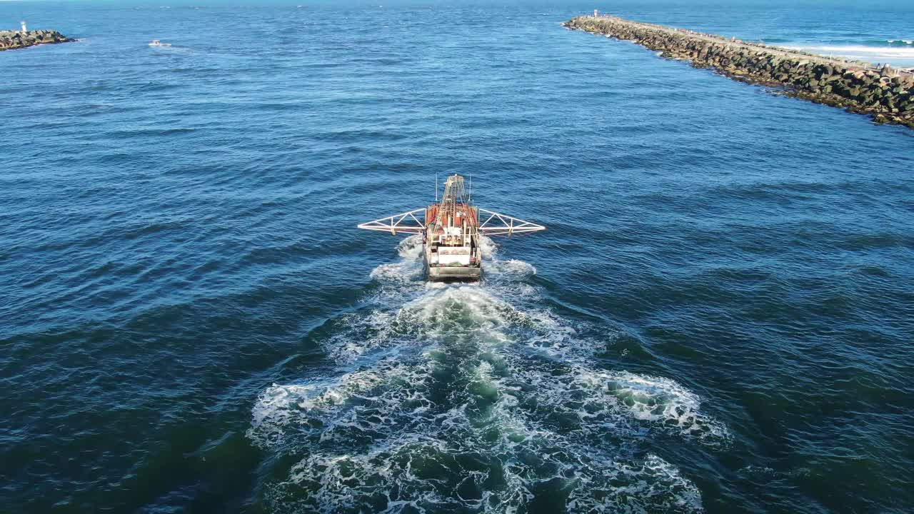 Prawn trawler leaving to fish from harbor sunset past rock wall.Drone