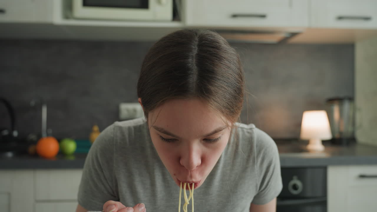 Jovem mulher branca comendo macarrão em cozinha moderna, inclinada sobre a tigela com vapor subindo, brilho da lâmpada e frutas no balcão, camisa casual, close-up espontâneo, estudante fazendo um lanche noturno, relaxada