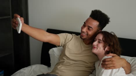 un feliz hombre moreno de piel negra en una camiseta crema se acuesta en la cama con su joven novia morena adulta con un peinado de bob en una camisa blanca y toma una selfie usando un teléfono blanco