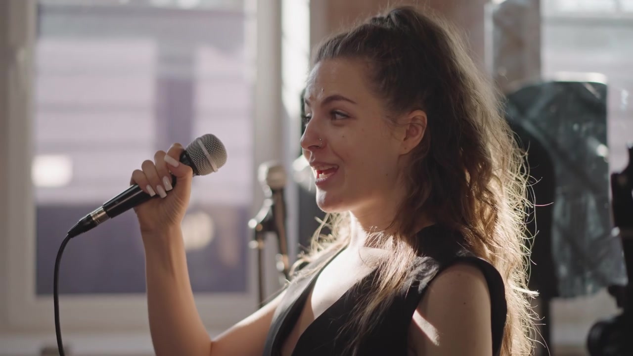 Young woman rehearsing with studio microphone, adjusting stand and warming up vocals in sunlit practice room, focused expression, music school atmosphere with equipment in background, musician