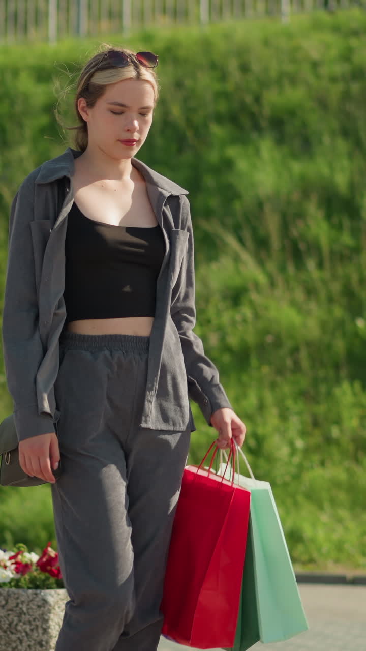 한 여인이 다채로운 쇼핑백을 들고 벤치에 다가와 앉아서 왼손을 벤치 위에 놓고 바람이 그녀의 머리카락과 초록색과 꽃을 흔들면서 자신을 조정합니다.