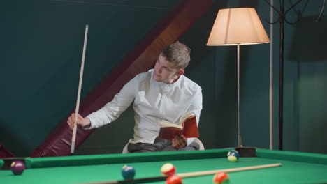 man in white shirt sits on white chair reading red book while holding cue stick near green pool table with scattered billiard balls. warm lighting from floor lamp creates relaxed atmosphere