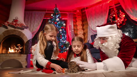 kids and santa during new year eve. girls and santa clause playing with little kitten on the background of fireplace and christmas tree. cozy atmosphere at christmas.