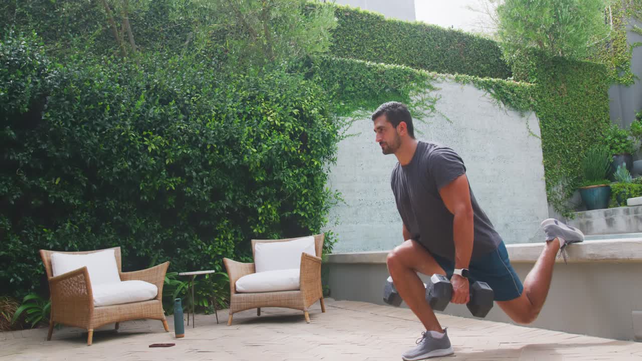 Uomo trentenne con il piede destro appoggiato al muro che esegue split squat con manubri sul patio, abbassandosi, mantenendo la posizione e risalendo per la potenza