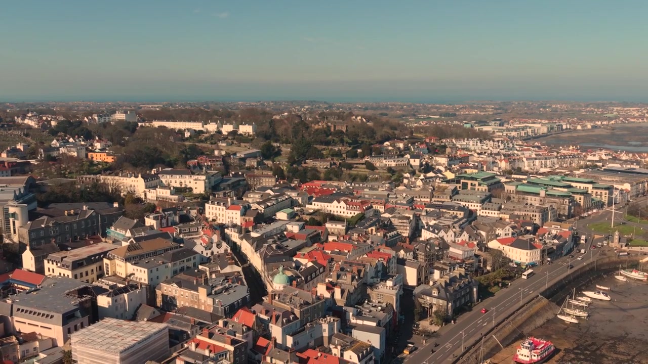 St Peter Port Guernsey Kanaløyene. Gradvis avdekking med høy droneflyvning over byen til sjøfronten i ettermiddagssolen, med utsikt mot Belle Greve-bukten og over hele øya under klar himmel.