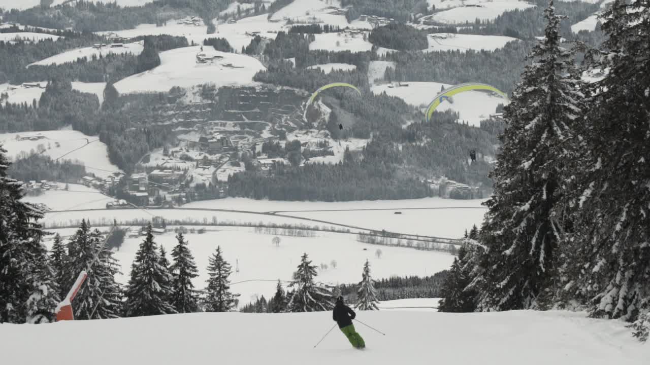 Inquadratura ampia di uno sciatore che scende una montagna mentre due parapendii planano in lontananza.