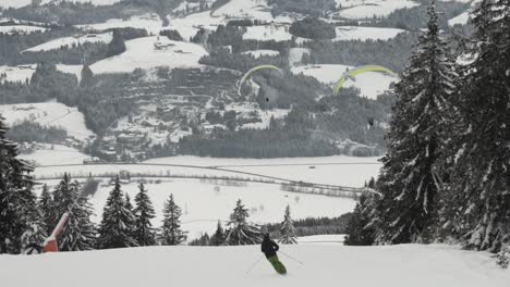 Weitwinkelaufnahme-Eines-Skifahrers,-Der-Einen-Berg-Hinunterfährt,-Während-Zwei-Gleitschirme-In-Der-Ferne-Gleiten