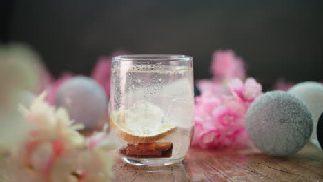 Gin-Tonic-Burbujeante-Con-Bloques-De-Hielo-En-Un-Vaso,-Hermoso-Video-De-Producto-De-Salón-De-Verano-Ligero-Con-Flores-Rosas-Y-Luces-Cálidas-Sutiles