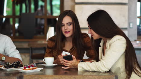 Two-girls-sitting-in-a-cafe-looking-at-the-phone-and-discuss-what-they-saw.-A-guy-comes-up-and-shows-a-funny-picture-on-his-teelphone