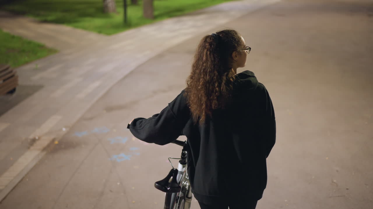Woman Riding Bicycle At Night, Contemplative Woman Guides Bicycle Along Peaceful Park Pathway At Night, Thoughtful Woman With Curly Hair Navigates Her Bicycle Along Quiet Park Trail After Sunset