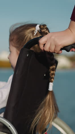 blonde preschooler girl sits in wheelchair. child frowns from bright sun looking at river on summer day on blurred background closeup backside view