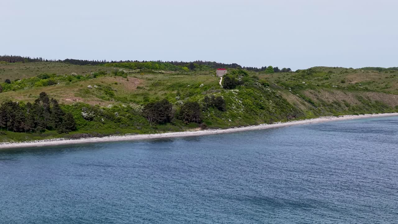 Drone footage shows a broad body of blue water alongside a grassy, green shoreline in Denmark. The calm water and horizon create a tranquil coastal landscape captured from above