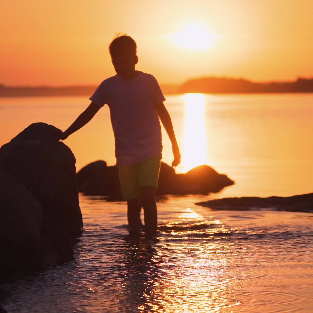 Boy walking in water at sunset. Silhouette of a child in clothes standing inside the river among stones. Kid splashing in the evening water. Slow motion.