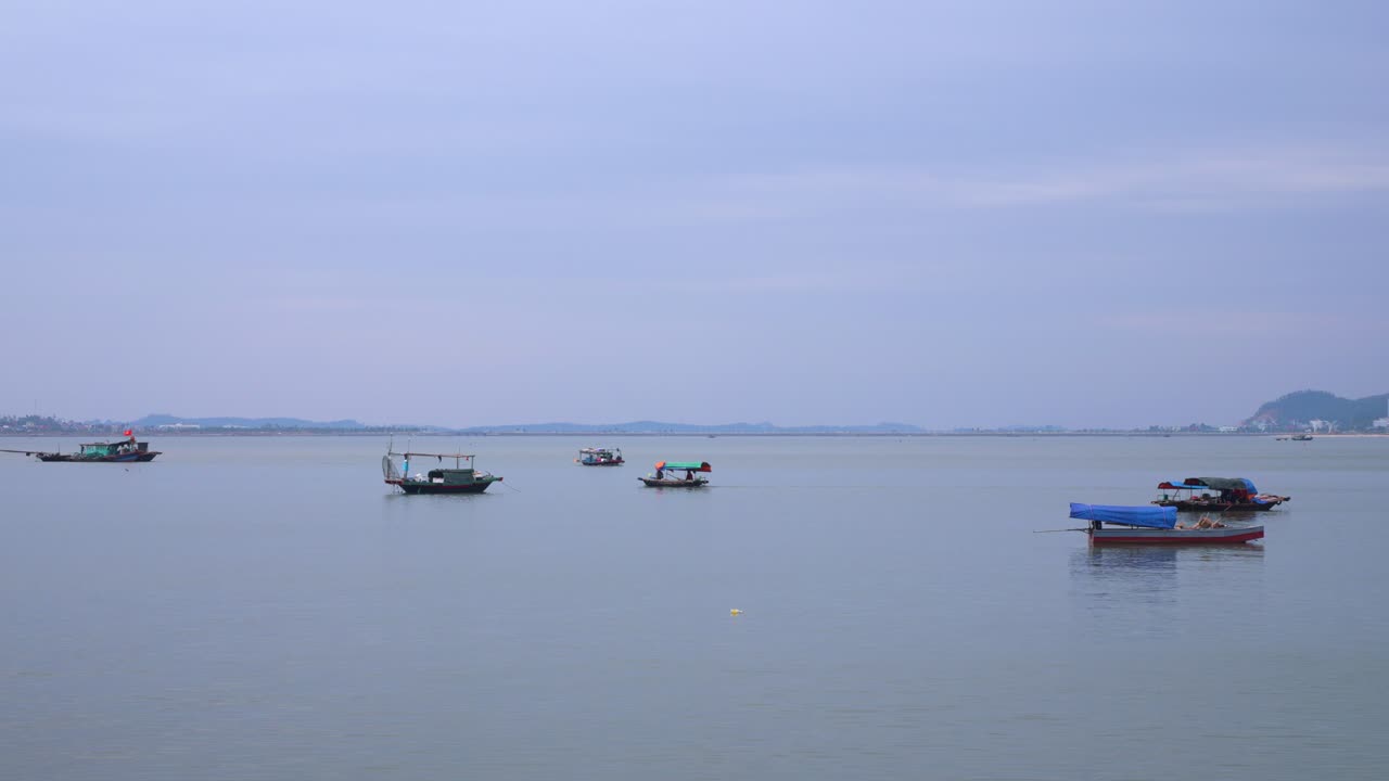 Several fishing boats float peacefully on the calm waters of Ha Long Bay under a misty lavender sky, with distant mountains creating a serene backdrop that showcases Vietnam's iconic seascape