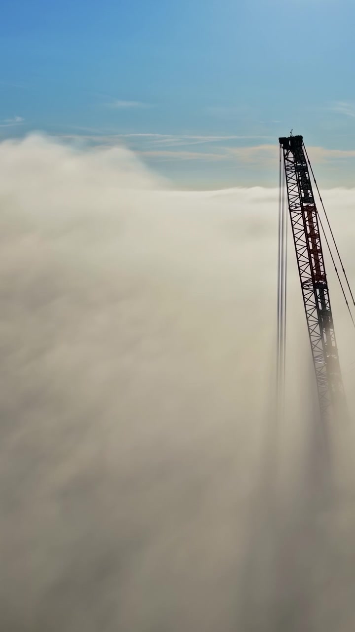 Gru edile che emerge sopra nuvole dense contro il cielo blu, sagoma verticale ripresa da drone