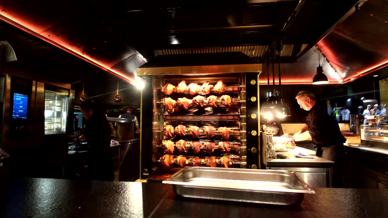 A night view through the window reveals chickens grilling, while staff inside prepare orders for the bustling crowd gathered outside in Munich.