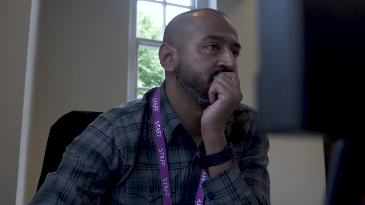 A close up shot from behind an office computer as an Asian Indian man sits with a clutched hand covering his mouth while reading the content displayed on a desktop monitor