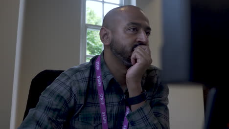 a close up shot from behind an office computer as an asian indian man sits with a clutched hand covering his mouth while reading the content displayed on a desktop monitor