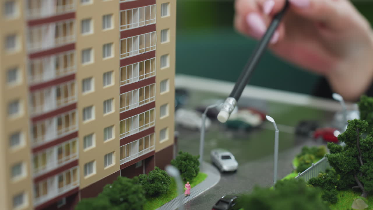 Close up of individual holding pencil while explaining architectural miniature of residential building surrounded by street layout, parked cars, and green landscaping during project presentation