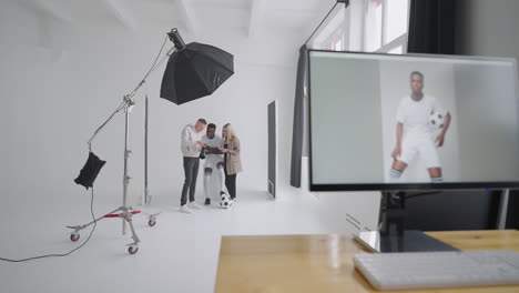 monitor with an advertising cover of the picture on the background a photographer a football player a model and a director look at photos from a photo shoot for the cover on a camera