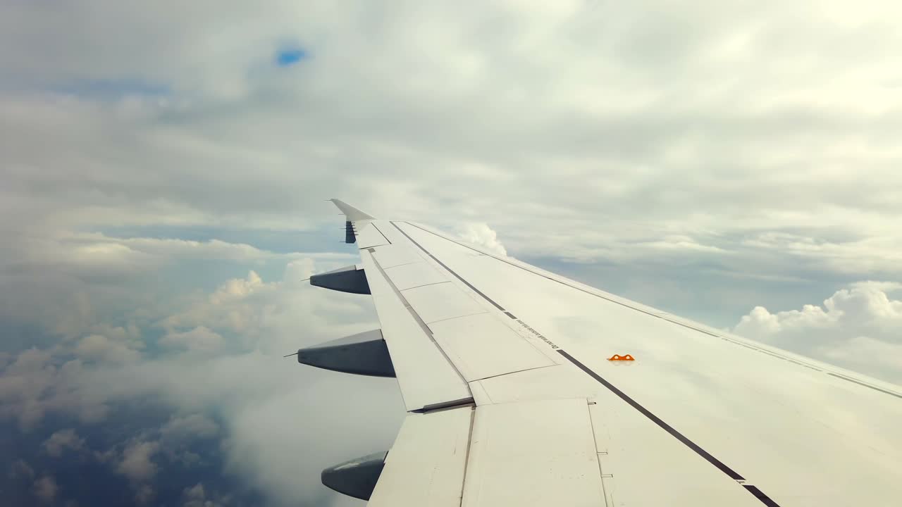 Watch as the airplane wing glides through fluffy clouds under a blue sky, showcasing breathtaking aerial views of the landscape below. Experience the serenity of flying