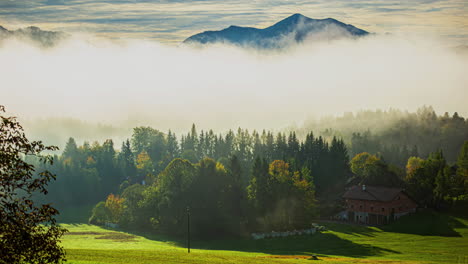 Timelapse-De-Un-Idílico-Paisaje-Montañoso-En-Austria-Mientras-Majestuosos-Picos-Montañosos-Se-Asoman-En-El-Horizonte-Y-La-Niebla-Se-Eleva-Pintorescamente-Sobre-El-Valle-Con-Vistas-A-Una-Granja-Al-Borde-Del-Bosque