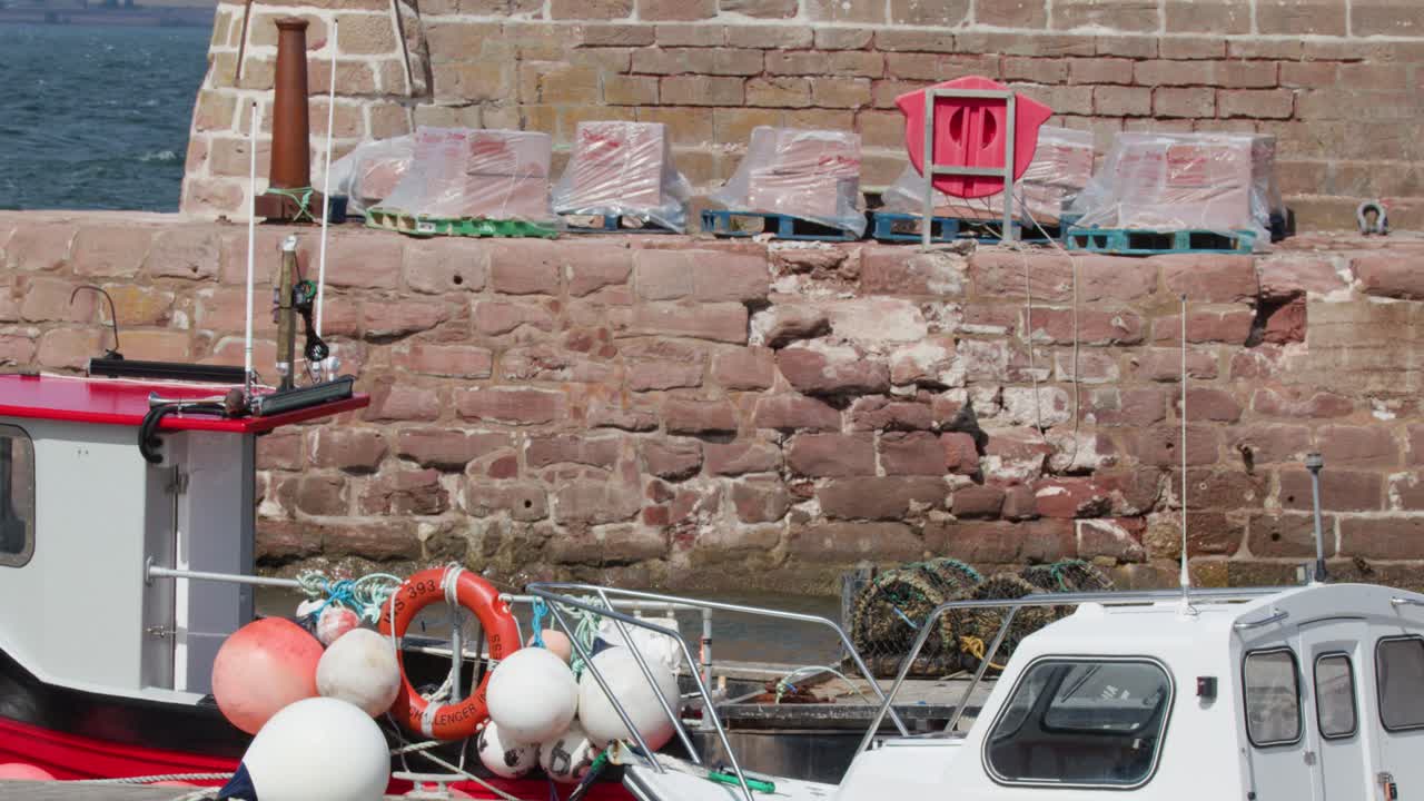Red fishing boat moves past moored vessel, colorful buoys, and stone harbor wall in daylight