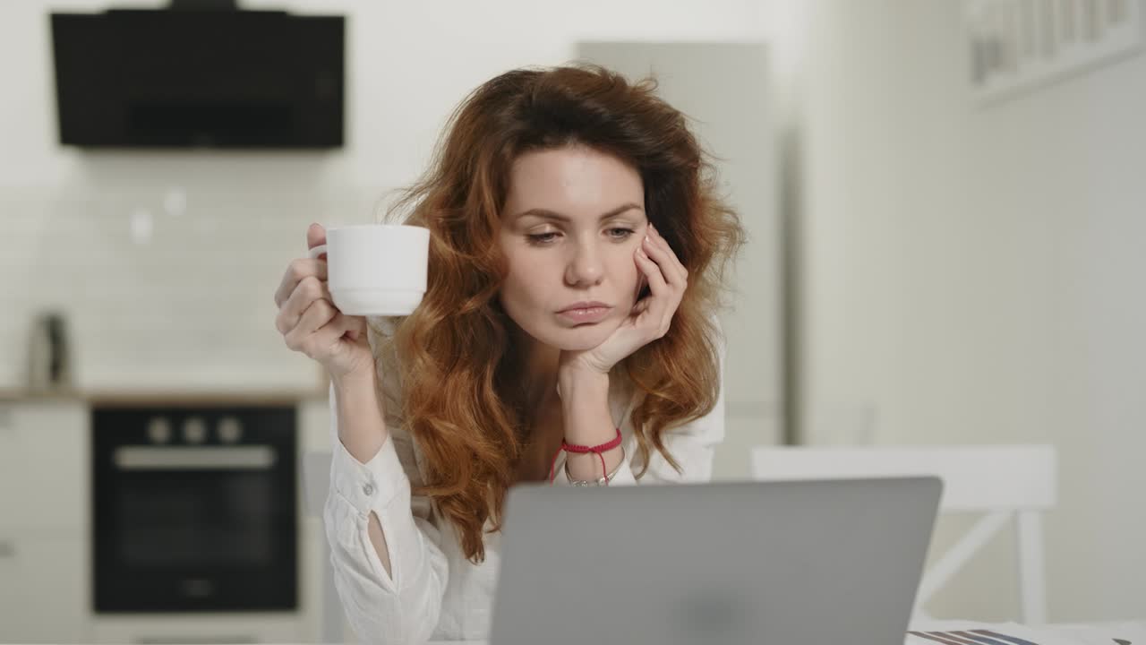 Concentrated woman drinking tea at morning kitchen. Young lady working laptop