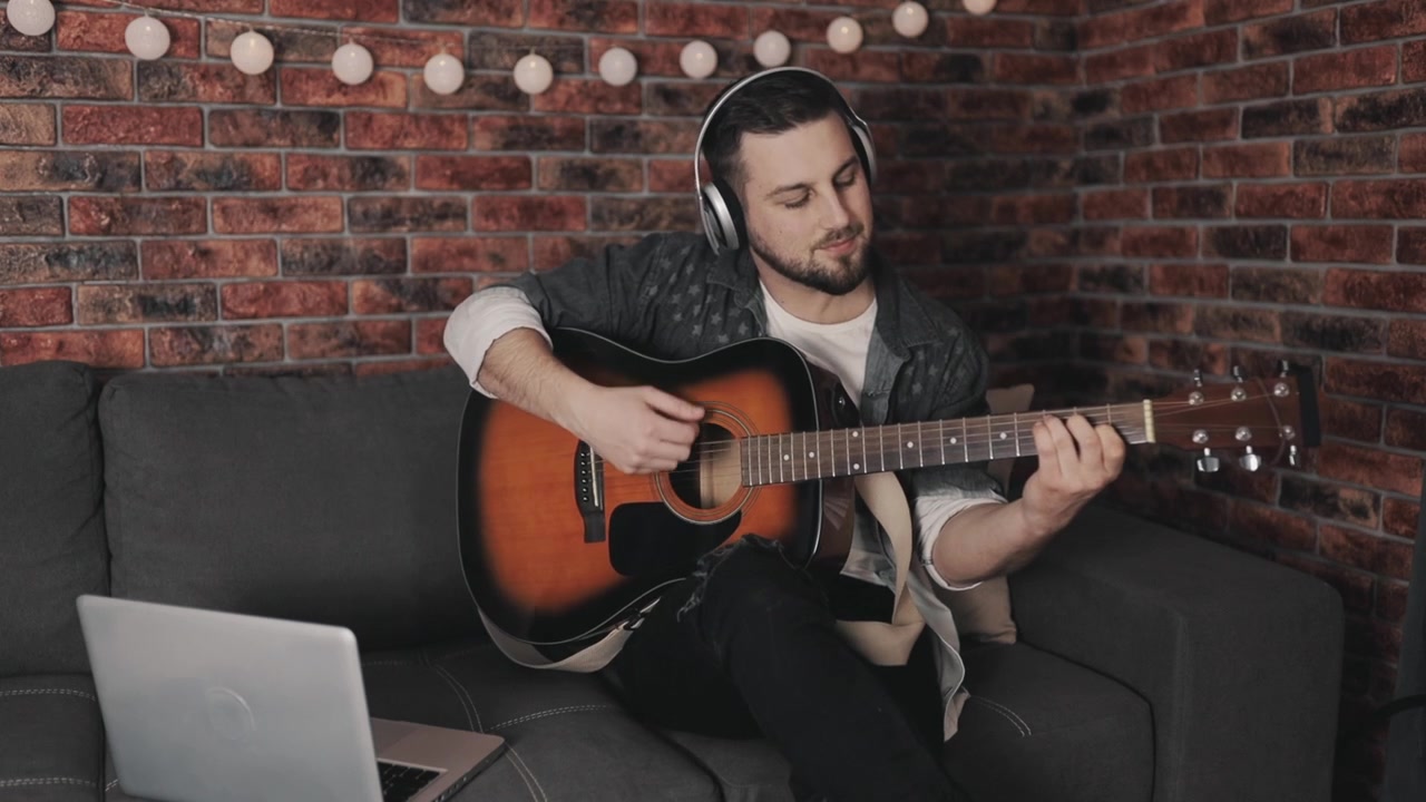 Ung mandlig musiker spiller guitar og bruger bærbar computer derhjemme