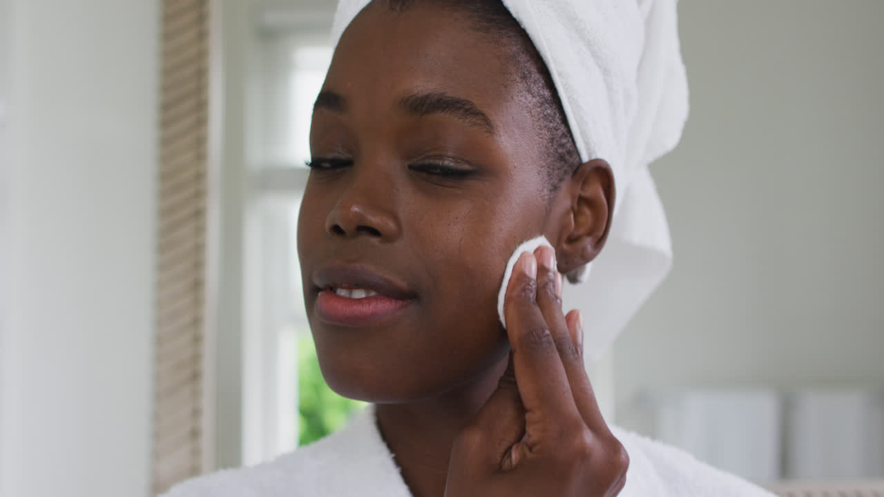 Portrait of african american woman in bathrobe cleaning her skin with a cotton pad while looking in 