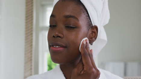 Portrait-of-african-american-woman-in-bathrobe-cleaning-her-skin-with-a-cotton-pad-while-looking-in-