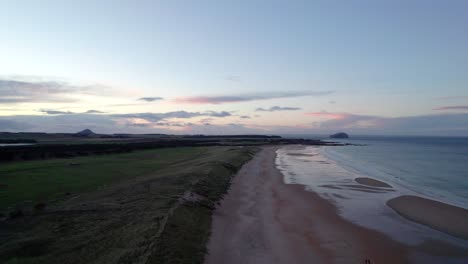 Drohnenaufnahmen,-Die-Während-Eines-Rosafarbenen-Sonnenuntergangs-Langsam-über-Einen-Langen-Sandstrand-Und-Sanddünen-Fliegen,-Während-Die-Flut-Sanft-Das-Ufer-Umspült-Und-Menschen-Am-Strand-Entlang-Gehen