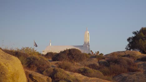 típica iglesia griega blanca con bandera griega ondeando en el viento con arbustos en primer plano contra el cielo azul