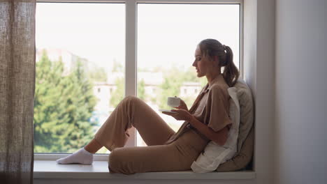 Woman-has-cup-of-hot-drink-enjoying-view-outside-window