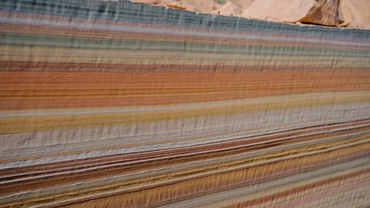 Static close view of striped sandstone layers in desert landscape, showing cream, tan, rust, green, and gray mineral bands, blurred cliffs behind, bright sunlight, and strong geological texture
