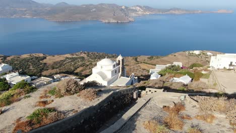 Drone-view-in-Greece-flying-over-a-white-church-on-a-hill-with-a-greek-white-house-town-facing-blue-sea-on-a-mountain