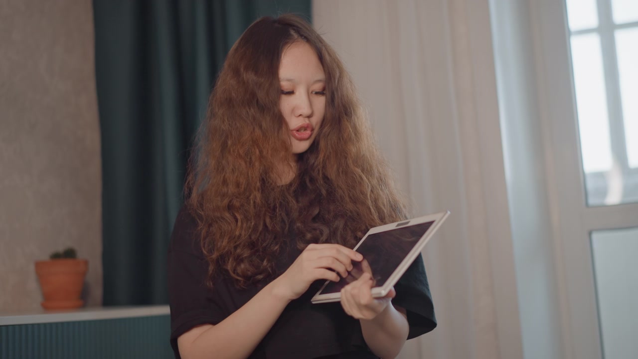 Young woman with long curly hair adjusts small blank chalkboard at chest level while looking down with focused, engaged expression in softly lit home interior with warm natural window light