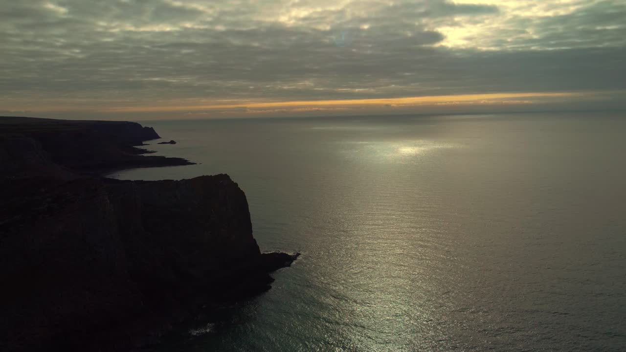 Panning Aerial View of Welsh Coastline with Bright Sun Reflecting on Ocean Behind Dark Clouds with Dynamic Rugged Coastline