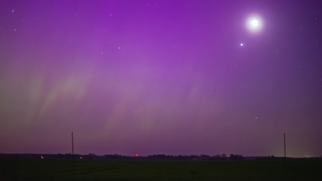 a bright moon stands high in a purple sky in which the green northern lights dance