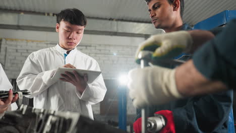 mechanic demonstrates tool usage on engine while students take notes on tablets, second mechanic observes student's input, industrial automotive workshop filled with tools