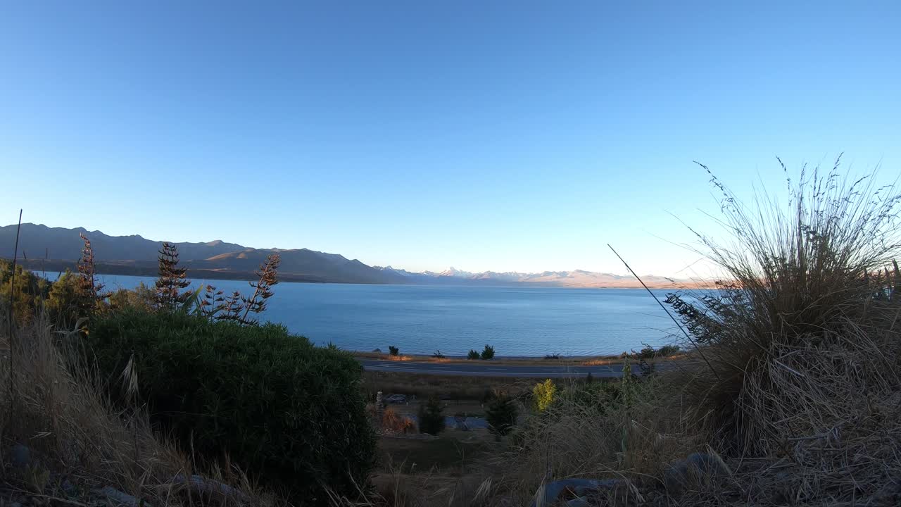 Day-to-night timelapse of Lake Pukaki with Mount Cook in the distance, foreground bushes and Highway 8 in frame as sunset fades to twilight