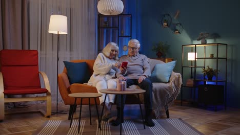 grandfather and grandmother talking making online shopping on mobile phone sitting at home sofa